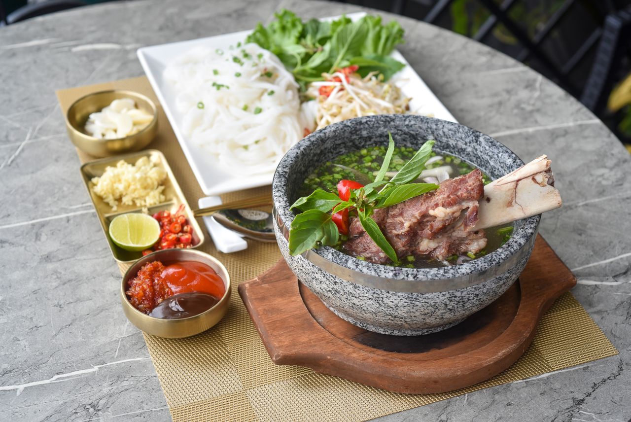 Stone bowl of Vietnamese Pho with beef bone, noodles, and fresh herbs on a table.
